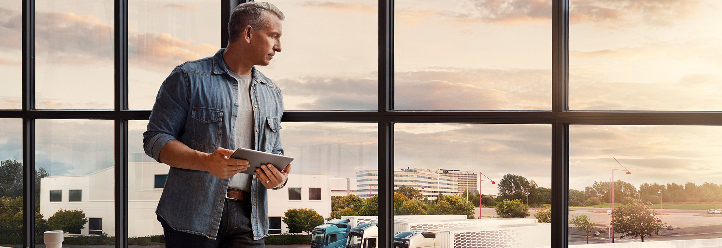 Мужчина с планшетом стоит у окна и смотрит на свой парк грузовых автомобилей