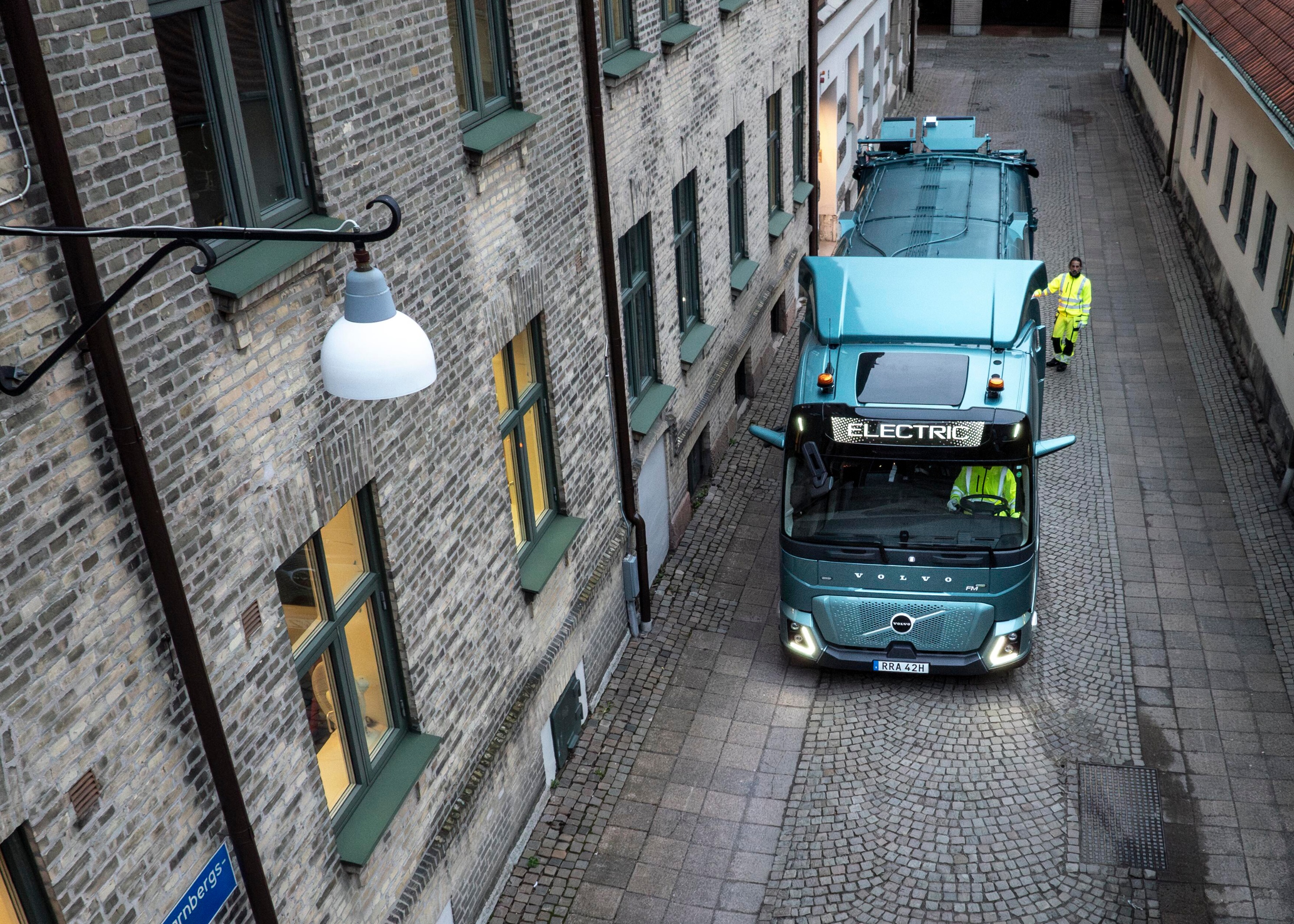 Volvo truck moving along a narrow cobbled city street beside brick buildings, with a driver and a worker in high-visibility clothing.
